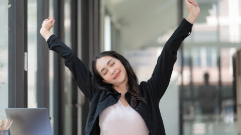 Taking a micro break at work. Woman raising arms at desk to stretch.